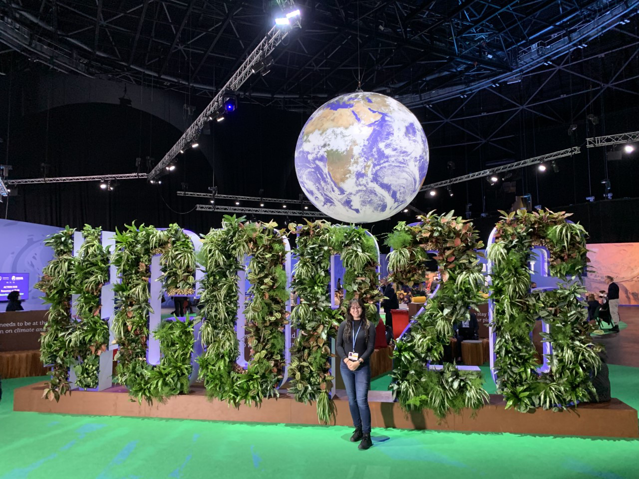 A young woman stands in front of an earth-themed installation that spells out "#COP26"